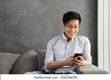 Portrait Of A Happy Young Asian Man Using Mobile Phone While Sitting On A Couch At Home