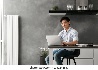 Portrait Of A Happy Young Asian Man Working On Laptop Computer While Sitting At The Table With Paperwork At Home