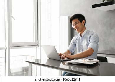 Portrait Of A Happy Young Asian Man Using Laptop Computer While Sitting At The Table With Paperwork At Home