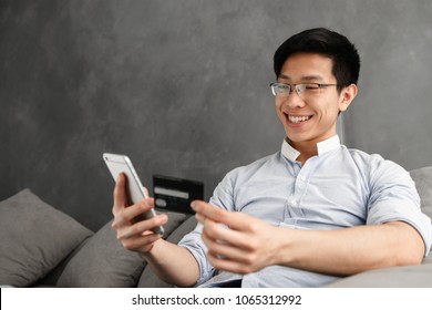 Portrait Of A Happy Young Asian Man Using Mobile Phone With Credit Card While Sitting On A Couch At Home