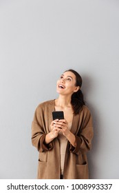 Portrait Of A Happy Young Asian Woman Holding Mobile Phone While Standing And Looking Up At Copy Space Over Gray Background