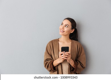 Portrait of a happy young asian woman holding mobile phone while standing and looking away over gray background