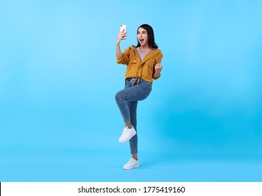 Portrait Of A Happy Young Asian Woman Celebrating With Mobile Phone Isolated Over Blue Background.