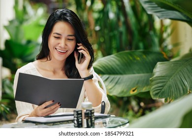 Portrait Of Happy Young Asian Woman Reading Catalog, Talking On Phone And Ordering Ingredients For Her Cosmetics Brand