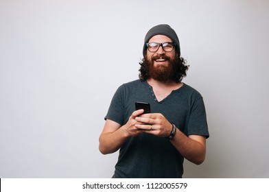 Portrait Of A Happy Young Bearded Hipster Man Holding Mobile Phone While Standing And Looking At Camera Over White Background