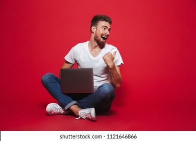 Portrait of a happy young bearded man pointing at copy space while sitting with laptop computer isolated over red background