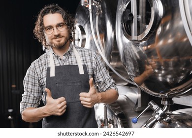 Portrait happy young bearded man shows thumbs up on background craft beer tank. Brewery worker with industrial equipment. - Powered by Shutterstock