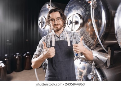 Portrait happy young bearded man shows thumbs up on background craft beer tank. Brewery worker with industrial equipment. - Powered by Shutterstock