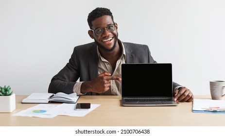 Portrait of happy young black businessman sitting at desk, pointing at laptop computer with blank black screen at home office, mockup. People, technologies and remote work concept