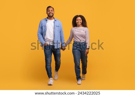 Portrait of happy young black couple holding hands and walking towards camera together, african american man and woman posing on yellow studio background, full length shot with copy space