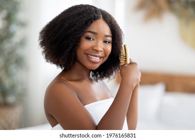 Portrait Of Happy Young Black Lady Brushing Her Curly Dark Hair At Home. Attractive African American Woman Taking Care Of Herself, Making Hairstyle, Using Wooden Brush, Smiling At Camera