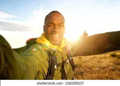 Portrait Of Happy Young Black Man Hiking With Backpack Taking Selfie And Pointing To Sunset