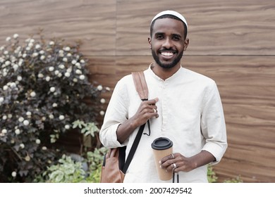 Portrait Of Happy Young Black Student Guy In Kufi Cap Holding Handle Of Satchel And Coffee Cup Outdoors
