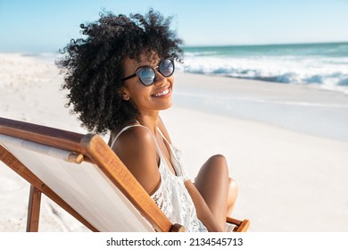 Portrait of happy young black woman relaxing on wooden deck chair at tropical beach while looking at camera wearing spectacles. Smiling african american girl with fashion sunglasses enjoying vacation. - Shutterstock ID 2134545713