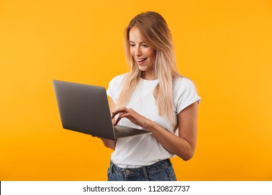Portrait of a happy young blonde girl using laptop computer isolated over yellow background