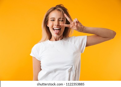 Portrait of a happy young blonde girl showing peace gesture while jumping and sticking her tongue out isolated over yellow background