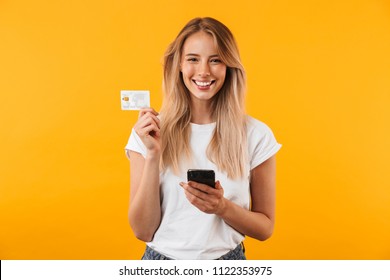 Portrait of a happy young blonde girl showing plastic credit card while holding mobile phone isolated over yellow background - Shutterstock ID 1122353975