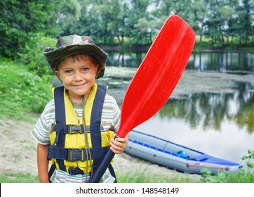 Portrait of happy young boy holding paddle near a kayak on the river, enjoying a lovely summer day