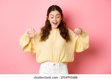 Portrait Of Happy Young Brunette Woman Checking Out Promo Offer, Pointing And Looking Down With Amazed Face, Standing Over Pink Background