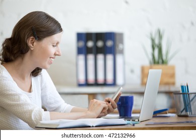 Portrait Of Happy Young Business Woman Working In Home Office Interior In Loft Holding Smartphone And Looking At Screen With Smile. Cheerful Office Person Using Mobile Phone, Typing, Texting Message