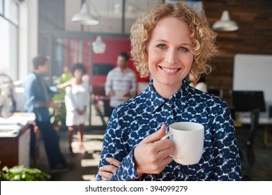 Portrait of happy young businesswoman holding a cup of coffee looking at camera and smiling. She is standing in her office with team of colleagues talking in background.