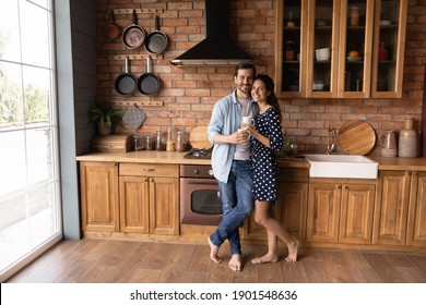 Portrait Of Happy Young Caucasian Couple Renters Or Tenants Pose In Cozy Modern Renovated Design Kitchen At Home. Smiling Millennial Man And Woman Hug Embrace Enjoy Family Morning Drinking Coffee.