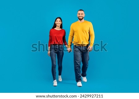 Portrait of happy young caucasian couple holding hands and walking towards camera together, man and woman posing isolated on blue studio background, full length shot with copy space
