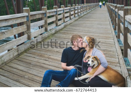 Portrait of happy young Caucasian couple with dog siting on wooden bridge and kissing