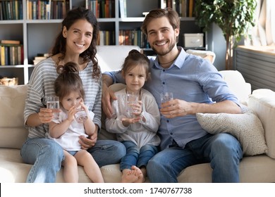 Portrait Of Happy Young Caucasian Family With Little Daughters On Couch At Home Recommend Drinking Water, Smiling Parents With Small Girls Children Follow Healthy Lifestyle, Dieting, Wellness Concept