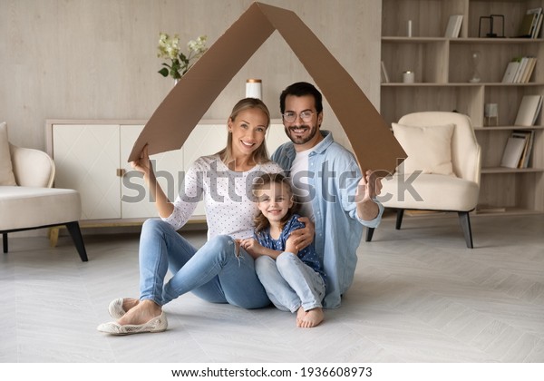 Portrait of happy young Caucasian family with\
daughter relax under roof celebrate relocation to own home. Smiling\
parents with girl child enjoy new house or apartment. Rental, real\
estate concept.