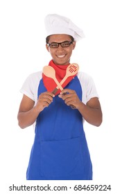 Portrait of a happy young chef holding two wooden spoons, guy wearing  blue chef uniform and chef hat with glasses, isolated on white background