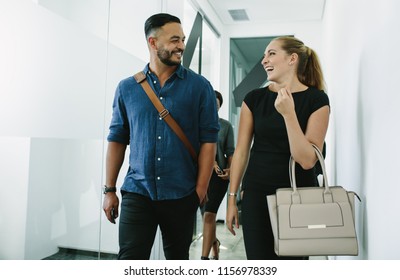 Portrait Of Happy Young Colleagues Leaving Office After Work. Young Man And Woman Walking Through Corridor Talking And Smiling.