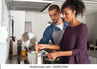 Portrait Of Happy Young Couple Cooking Together In Kitchen At Home. Woman Cooking In Pot With Her Boyfriend In Kitchen. Man In Apron Helping His Girlfriend Preparing Dinner.