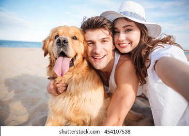 Portrait Of Happy Young Couple Hugging Their Dog And Smiling On The Beach