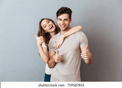 Portrait Of A Happy Young Couple Hugging While Standing And Showing Thumbs Up Gesture Over Gray Wall