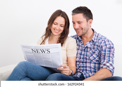 Portrait Of A Happy Young Couple Sitting On Couch Reading Newspaper