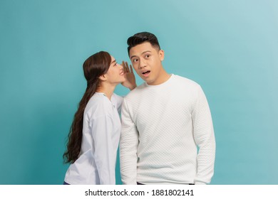 Portrait of a happy young couple standing together over blue background, telling secrets