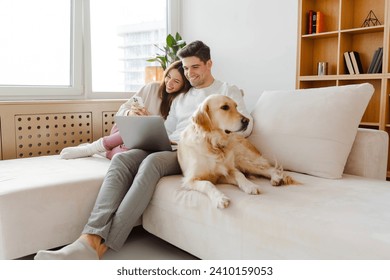 Portrait of happy young couple using laptop, man holding mobile phone, hugging dog, sitting together on comfortable sofa at home. Online shopping concept - Powered by Shutterstock