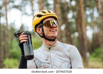 Portrait Of A Happy Young Cyclist In A Helmet And Wearing Glasses With A Bottle Of Water In His Hands On A Background Of Forest, Looking Away And Smiling.