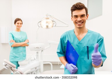 Portrait of a happy young doctor dentist in protection gloves, in the background a woman assistant holding documents and recording of data, in the dental clinic