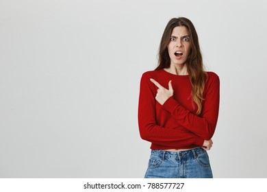 Portrait Of Happy Young European Good-looking Girl With Long Dark Hair In Casual Outfit Pointing Aside With Angry Expression Being Mad After Friend Said Offensive Joke About Her Weight.