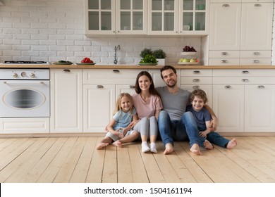 Portrait of happy young family couple sitting on wooden floor at kitchen with cute little kids siblings. Smiling positive father mother and small children looking at camera, enjoying weekend time. - Shutterstock ID 1504161194