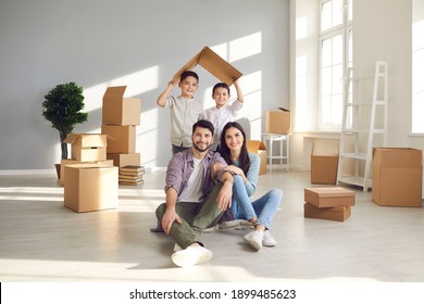 Portrait of happy young family in new home. Smiling mom and dad sitting on floor with unpacked boxes, and children holding symbolic roof behind. Real estate, mortgage, buying and moving house concept