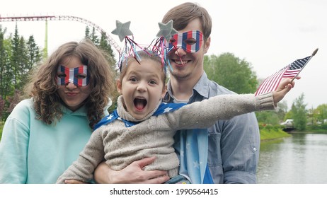 Portrait Of Happy Young Family Standing Outside In A Park During Independence Day, Looking At The Camera And Having Fun Making Silly Faces, Waving Usa Flags. Front View. 4th July Celebration.