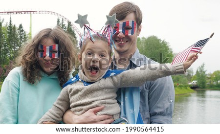Portrait of happy young family standing outside in a park during Independence Day, looking at the camera and having fun making silly faces, waving usa flags. Front view. 4th July celebration.
