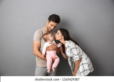 Portrait Of A Happy Young Family With Their Little Baby Girl Isolated Over Gray Background, Kissing