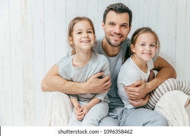 Portrait Of Happy Young Father Embraces His Two Daughters, Loves Them Very Much, Pose Together At Home. Single Dad With Beautiful Femle Children Have Cheerful Expressions. Fatherhood Concept