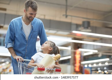 Portrait of happy young father grocery shopping in supermarket and smiling at little girl sitting in shopping cart, copy space