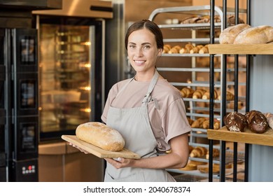 Portrait of happy young female baker in apron standing with fresh loaf of bread by workplace - Shutterstock ID 2040678374