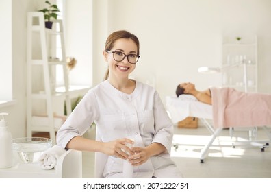 Portrait of happy young female dermatologist, skin therapist, beautician and skincare professional at work. Beautiful woman in clean white coat uniform and glasses sitting in her office and smiling